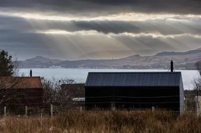 a building on the side of a field with a body of water at Jura - The Black Hut in Isle of Jura
