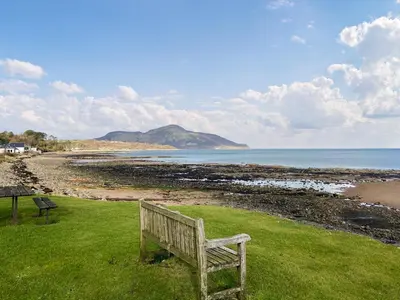 a wooden bench sitting on the grass near the beach at Cosy Cottage in Lamlash