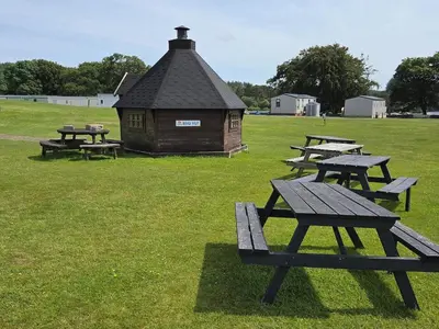 a group of picnic tables in a field with a building at Lovely Caravan number 96 at Three Lochs Holiday Park in Kirkcowan