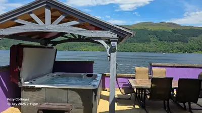 a hot tub on a deck with a table and water at Jenny's Bay with Hot Tub in Lochgoilhead