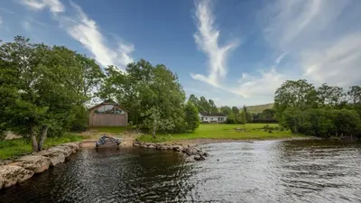 a river with a barn and a house in the background at Ardeonaig Lodge with Hot Tub & Water Fronted Games room in Ardeonaig