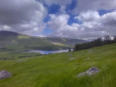 a green hill with a lake in the middle of a field at Holiday Home Benmore Farm House in Stirling mit Bergblick by Interhome in Crianlarich