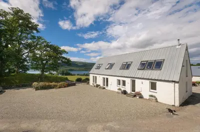 a white barn with a gravel driveway next to a lake at Blarghour Farm Cottages in Ardchonnell