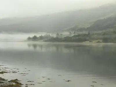 a misty lake with trees and a hill in the background at The Boathouse in Glencoe