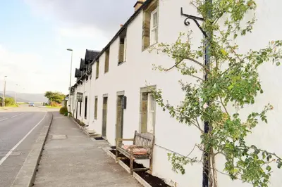 a white building with a bench next to a street at Newton Cottage North in Inveraray