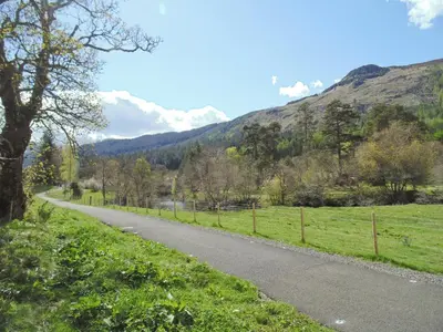 a road in the middle of a field with a mountain at Craigdarroch Cottage in Strathyre