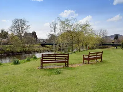 two benches sitting in the grass next to a river at Caroch Cottage in Comrie