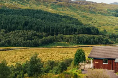 a small red house in a field next to a mountain at Kingfisher Lodge with sensational loch views in Crianlarich