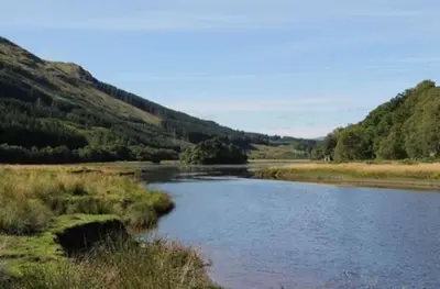 a river in the middle of a field with mountains at Osprey Lodge with river and mountain view in Crianlarich