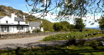 a white house with a stone wall next to a road at The Colonsay Hotel in Isle of Colonsay