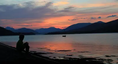 a person sitting on the shore of a lake at sunset at Camus Bhan in Ballachulish