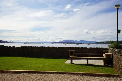 a wooden bench sitting in the grass near a body of water at Beach house no2 with amazing sea views and private garden onto beach. in Fairlie
