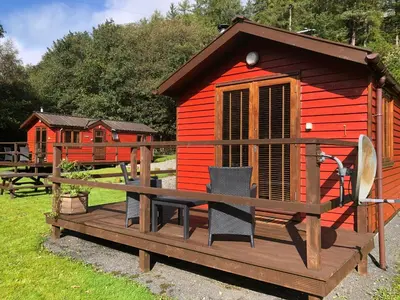 a red cabin with chairs on a wooden deck at Rashfield Sheilings - Quiet Riverside Lodges for Couples near Pucks Glen in Dunoon