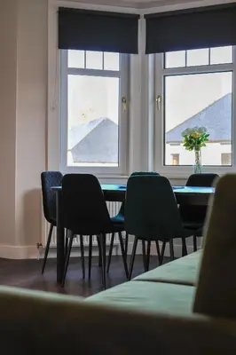 a dining room with a table and chairs and two windows at The White City Apartment in Oban