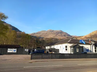 a building on the side of a road with mountains in the background at Riverside Pod, West Highland Way Holidays in Kinlochleven