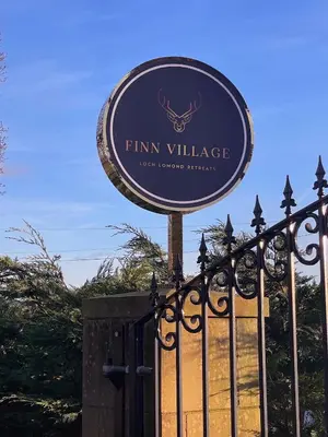 a sign for a firm village behind a fence at FINN VILLAGE Loch Lomond Villa - THE BLACK ROOM with Private Jacuzzi-Style Bath in Drymen
