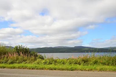 a road next to a lake with a cloudy sky at Chapelton Cottage in Strachur