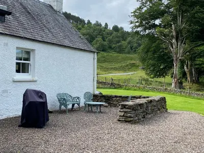 a house with two chairs and a bench next to a stone wall at Riverside Cottage, Bridge of Balgie, Glenlyon, Perthshire in Bridge of Balgie