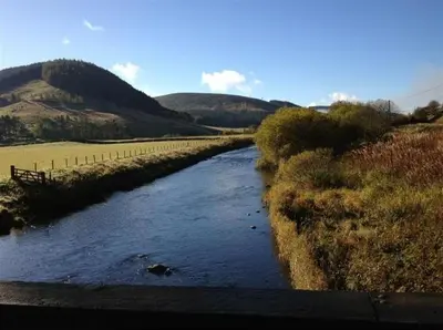 a river in the middle of a field with hills at Manyleith Rig Cottage in Polmood